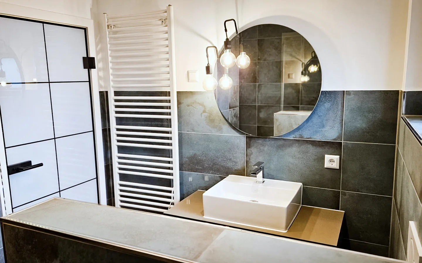 Badsanierung Schwetzingen – Badezimmeroase mit dunklem Fliesenspiegel, rechteckiges Waschbecken mit silberner Armatur, runder Eyecatcher Spiegel, milchige Glastür und Wandheizung mit Handtuchhalter-Funktion