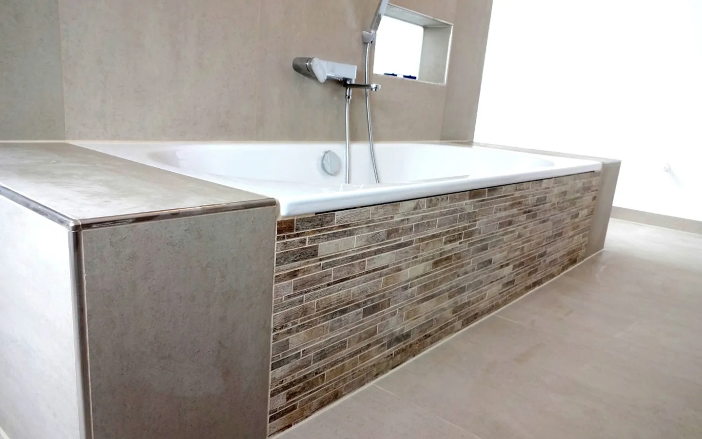 Bad umbauen lassen – neu von Sanitärfirma fertiggestelltes Badezimmer mit schönen Fliesen in Taupe-Ton mit edler Unterputz Badewannenarmatur und großer Badewanne mit passender Verkleidung aus Feinsteinzeug und Mosaikfliesen